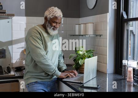 Senior African American man wearing light green sweater and jeans in kitchen using laptop and phone Stock Photo