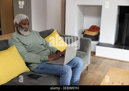 Senior African American man wearing sage sweatshirt sitting on sofa using laptop with phone nearby Stock Photo