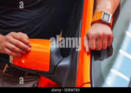 man wearing an orange digital watch and a black t-shirt has his hands resting on the side mirror and door frame of a bright orange car. Stock Photo