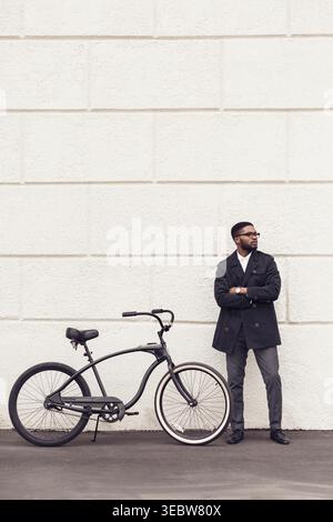 Man in Stylish Coat Stands Beside Bicycle Against Wall in Urban Environment Stock Photo