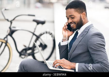 Businessman Working on Laptop While Talking on Phone Next to Bicycle in Urban Setting Stock Photo