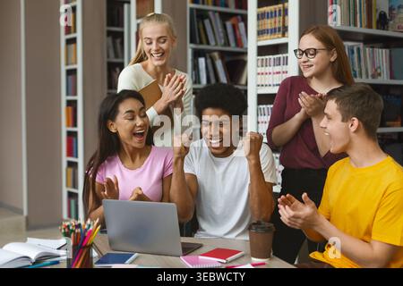 Group of Students Celebrating Success While Using a Laptop in a Bright Library Setting Stock Photo