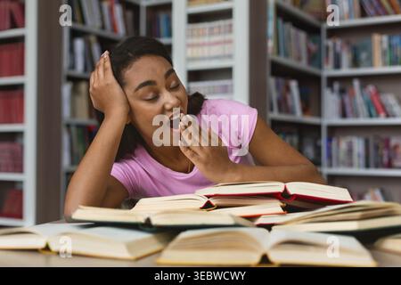 Young Woman Yawning While Studying Surrounded by Open Books in a Library Stock Photo