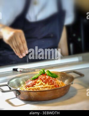 A professional chef in a dark blue apron adds a final touch of grated parmesan cheese to a dish of classic spaghetti bolognese garnished with fresh ba Stock Photo