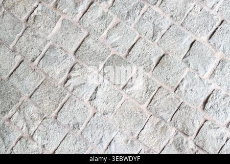 A top-down view of a grey cobblestone pavement featuring square-cut natural stones arranged in a diagonal pattern. The texture is rough and weathered, Stock Photo