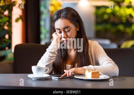 Young Woman Sitting Alone at a Cafe Table With Coffee and Cake While Using Her Phone Stock Photo
