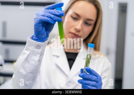 Laboratory technician analyzing green liquid specimen in graduated test tube while holding another with plant material. Clean lab environment with scientific equipment visible in background. Stock Photo