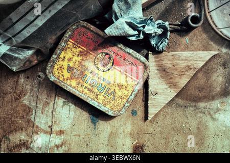 A small, rusty tobacco tin on a dirty wooden bench surrounded by wood, plastic and paper Stock Photo