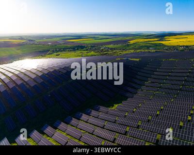 Large arrays of solar panels fill an expansive field in a rural area. The landscape shows green hills and blue skies. Sunlight reflects off the panels Stock Photo