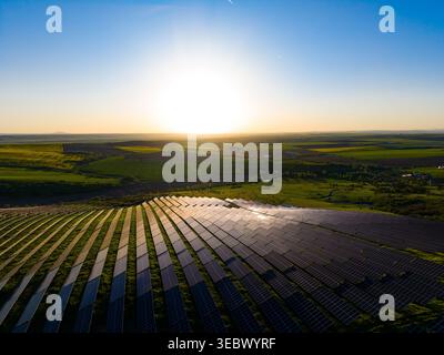 Sun sets in the background as solar panels cover a hillside. Fields stretch into the distance, highlighting the use of clean energy in a rural setting Stock Photo