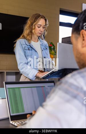 Coworkers working, standing woman in denim jacket using laptop riser at office spreadsheet visible Stock Photo