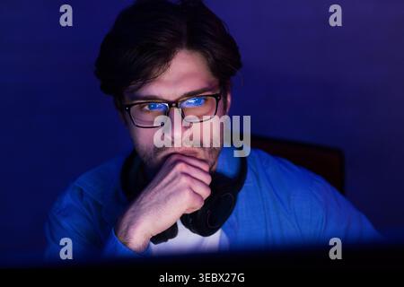 Young Man Wearing Glasses Deep in Thought While Working at Computer in Dimly Lit Room Stock Photo