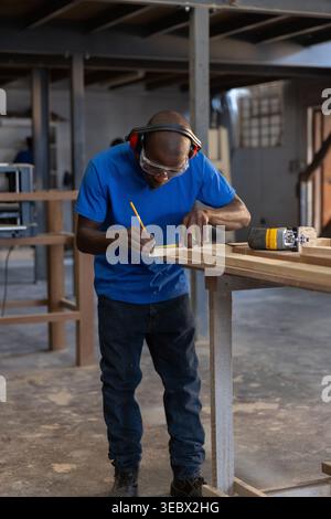 Long wooden board being measured and marked on workbench in carpentry workshop, with power router Stock Photo