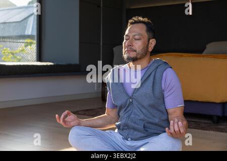 Middle-aged Asian man meditating cross-legged on floor in bedroom in hoodie, mustard throw nearby Stock Photo