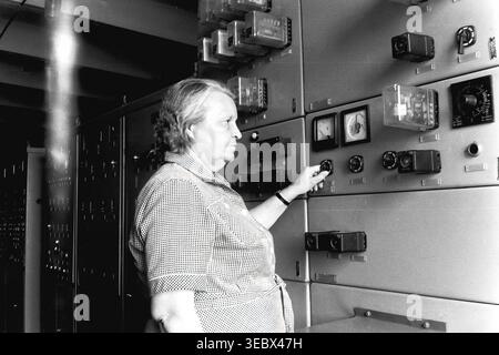 Slaviansk, 1986. At the traction power substation. An older female operator is adjusting switches on a large industrial electrical control panel covered with various analog meters, relays, and indicators. She is wearing a checkered short-sleeve shirt. The photograph captures the critical technical work required to maintain the city's trolleybus power supply during the Soviet era. This authentic archival image reflects the dedicated labor, complex infrastructure, and everyday industrial reality in the historical context of a peaceful Donbas. Stock Photo