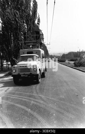 Sloviansk, 1986. Two technicians perform maintenance on the overhead contact network for the city's trolleybus system. The workers are elevated in the basket of a specialized GAZ utility truck parked on the side of a wide urban road. Wearing hard hats, they carefully inspect and repair the electrical lines ensuring the reliable operation of public transport. This historical documentary photograph highlights the daily industrial labor and infrastructure maintenance of the Soviet period, capturing the urban environment of a peaceful Donbas. Stock Photo