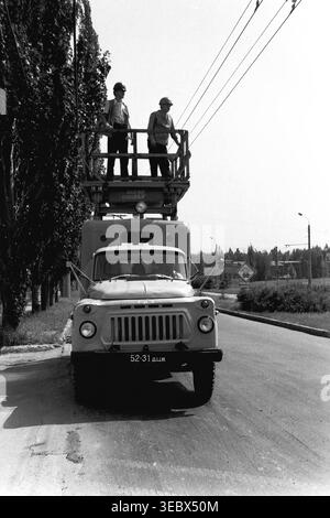 Sloviansk, 1986. Two technicians perform maintenance on the overhead contact network for the city's trolleybus system. The workers are elevated in the basket of a specialized GAZ utility truck parked on the side of a wide urban road. Wearing hard hats, they carefully inspect and repair the electrical lines ensuring the reliable operation of public transport. This historical documentary photograph highlights the daily industrial labor and infrastructure maintenance of the Soviet period, capturing the urban environment of a peaceful Donbas. Stock Photo