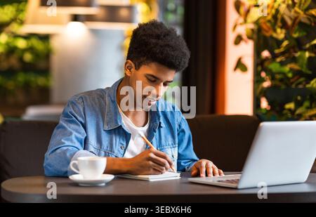 Young Man Writes Notes While Sitting at a Cafe Table With a Laptop and Coffee in a Cozy Environment Stock Photo