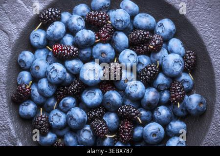 Close up view of blueberries and mulberries filling the black plate. Fresh blue berries texture, antioxidant superfood concept. Top view Stock Photo