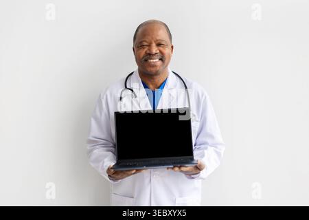 Smiling mature black doctor in white coat holding laptop with blank screen Stock Photo