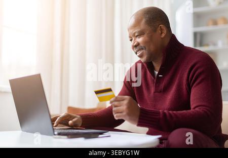 Happy mature black man holding credit card while using laptop at home Stock Photo