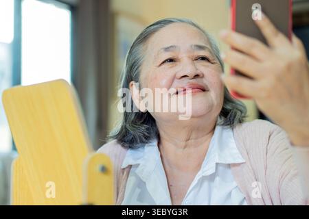 Happy senior Asian woman taking selfie in front of mirror at home. Selective focus. Smiling mature old lady pampering, healthy moisturized skin care c Stock Photo