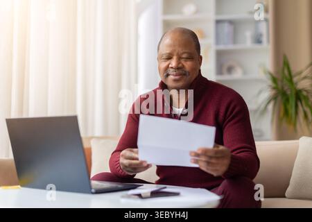 Pleased mature African American man reading paper document beside laptop at home Stock Photo