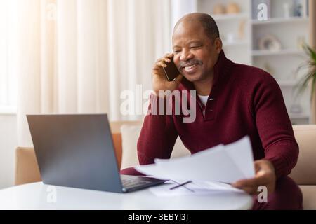 Smiling mature black man talking on smartphone and reviewing documents at home Stock Photo