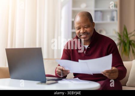Happy mature black man reviewing paper documents next to laptop at home Stock Photo