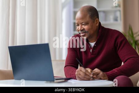 Smiling mature black man taking notes while working on laptop at home Stock Photo