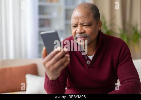 Confused mature black man squinting and frowning at smartphone screen at home Stock Photo