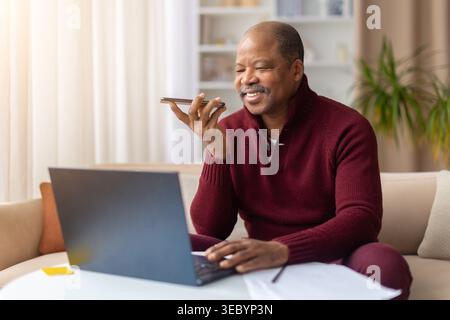 Black man using smartphone voice assistant while working on laptop at home Stock Photo