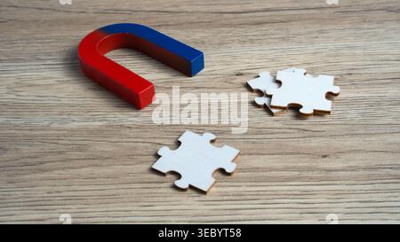 Large horseshoe magnet pulling white jigsaw puzzle pieces across a wooden surface. Concept for problem solving, logical attraction, and strategic solu Stock Photo