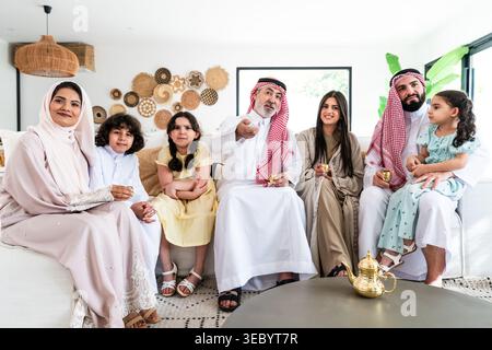 Arab family across three generations — grandparents, parents and children — sitting together in a modern living room, sharing traditional coffee, conv Stock Photo