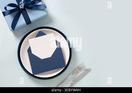 Blue gift box with ribbon envelope and blank card on plate with cutlery, minimal composition on light background, celebration concept, copy space. Fat Stock Photo