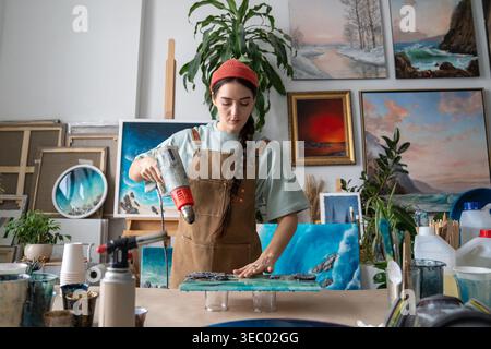 Concentrated woman designer working on epoxy resin painting at worktable in home art studio. Stock Photo