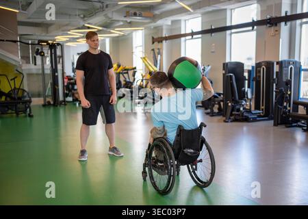 Disabled sportsman in wheelchair doing exercises with med ball on personal gym session with coach. Stock Photo