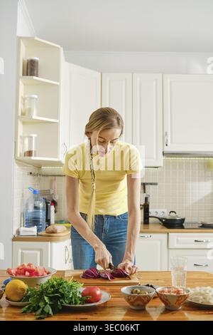 A person stands at a wooden table in a kitchen, chopping vegetables. There are bowls with ingredients nearby. The kitchen has white cabinets and a bri Stock Photo