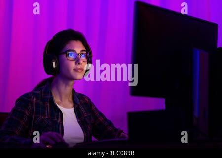 Young Woman Focused on Computer Screen in a Purple Lit Room at Night While Gaming Stock Photo