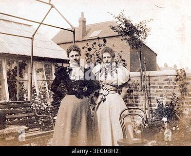 Two adults stand in a garden beside a wooden chair, with a brick wall and greenhouse behind them. Both wear high‑necked blouses, puffed sleeves, and long skirts. A house with a chimney rises above the wall, while climbing plants and a bench complete the scene. From a late Victorian album of small photographs taken by an amateur photographer. Most date between 1898 and 1901 and seem to relate to the family of John White (possibly of Sth London), a builder and contractor. The quality of the original photographs was variable and many were faded and discoloured. Stock Photo