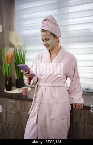 Woman wearing bathrobe and face mask checking her phone in the kitchen Stock Photo