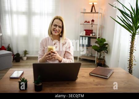Smiling woman enjoys coffee while working from home in cozy and stylish office space during daylight Stock Photo