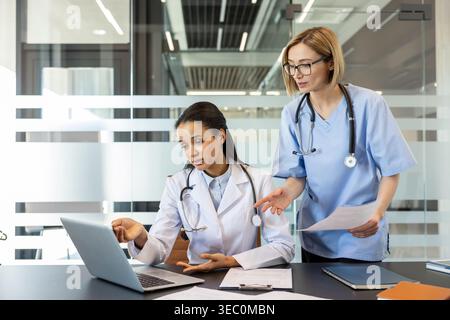 Two diverse female healthcare professionals review patient data on a laptop in a modern clinic, collaborating on diagnosis, treatment planning and tel Stock Photo