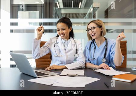 Two cheerful female doctors or nurses collaborating and celebrating successful work outcomes online while looking at a laptop in a modern clinic offic Stock Photo