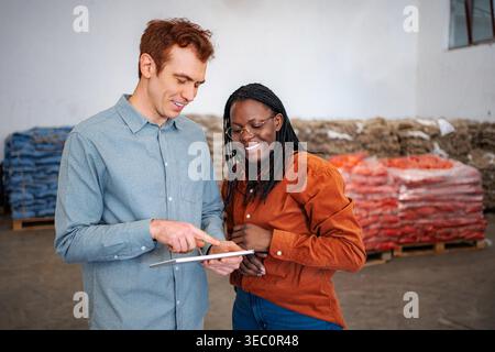 Two diverse workers smiling while checking inventory records on a digital tablet in a warehouse Stock Photo