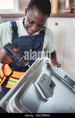 Smiling female technician repairing a dishwasher using an electric screwdriver in a kitchen Stock Photo