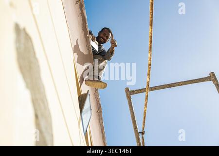 Marsa alam, egypt 21 april 2026: happy male construction worker on scaffolding giving thumbs up, smiling confidently against a clear blue sky backdrop Stock Photo
