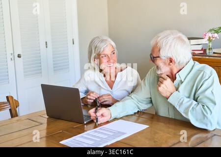 A senior couple is engaged in retirement planning and financial protection, reviewing important documents and using a laptop at their home. Stock Photo