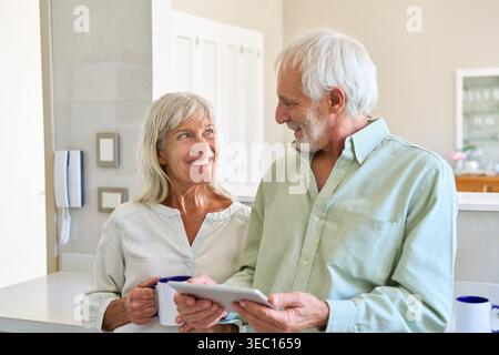 A senior couple is engaged in retirement planning and digital inclusion, using a tablet together in their bright, modern kitchen. Stock Photo
