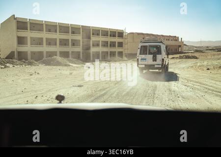 Marsa alam, egypt 21 april 2026: white suv driving on an unpaved road, leaving a dust trail past unfinished buildings in the arid marsa alam desert la Stock Photo
